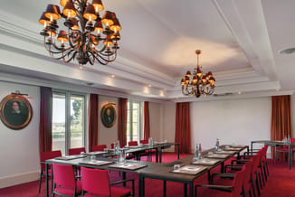 Moderne vergaderzaal met zwarte vergadertafels, rode stoelen en rode tapijt. Twee gouden kroonluchters aan plafond met casset