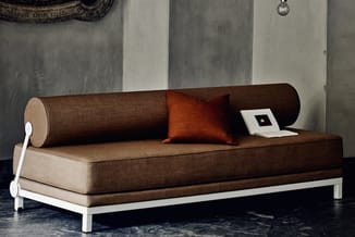 Bruine loungebank met cilindervormige rugkussen en oranje sierkussen, witte metalen frame, op donkere vloer in minimalistisch