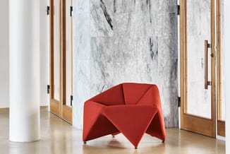 Rode sculptuurale fauteuil met gevouwen stoffen vorm op metalen poten in moderne kantoorruimte met marmeren wand.