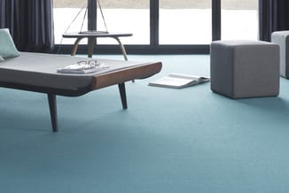 Licht kantoor met turquoise vloerbedekking, grijze bank, laag tafel met houten armsteun, grijze poef en grote ramen met grijz