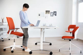 Vrouw aan staand bureau met rood-witte ergonomische bureaustoel, tweede rode stoel rechts, moderne kantoorruimte met witte wa