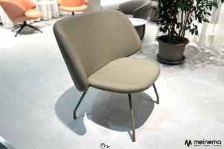Grijze stoel EVY met gestoffeerde zitting en rug, metalen poten, in showroom met plant en andere meubels.