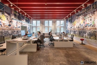 Modern kantoor met witte bureaus, zwarte bureaustoel, rode plafondbalken en historische fotowanden. Grote ramen, grijs tapijt