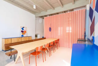 Vergaderruimte met lichte houten tafel, oranje kunststoffstoelen, houten zijkast met blauwe bovenkant, roze verticale raambek