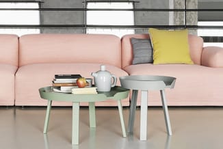 Roze bank met gele en grijze kussens, twee groene en grijze bijzettafels met boeken, appel en theepot in moderne kantoorruimt
