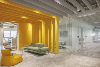 Modern kantoorruimte met gele verticale scheidingswanden, groene sofa, gele loungestoel, glazen kantoorwanden en beige vloer.