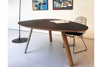 Ovale vergadertafel met zwart blad en houten poten, naast draadstoel en staande lamp in minimalistische kantoorruimte.