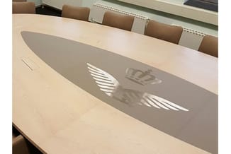 Grijze akoestische scheidingswand op lichte houten vergadertafel met witte logo's en vleugelmotieven.