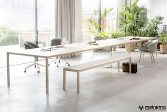 Modern kantoor met lichte houten vergadertafel, witte blad, bijpassende zitbank, grijze stoelen, veel groen en grote ramen me