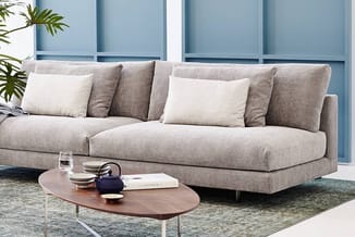 Grijze hoekbank met beige kussens in moderne kantoor-/woonruimte met houten salontafel en blauwe panelen op achtergrond.