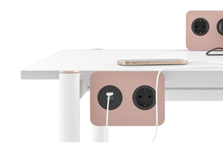 Roze powerbox met twee stopcontacten en witte kabel, gemonteerd onder een wit bureau met smartphone erop.