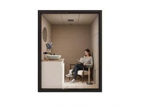 Kleine werkcel met witte wastafel, ronde spiegel, houten blad en zwarte kraan. Vrouw zit op houten stoel te lezen. Beige wand
