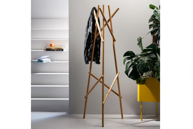 Houten garderobe met X-vormig design, beladen met donker kledingstuk, in kantoorruimte naast gele plant pot en grijze wandkas