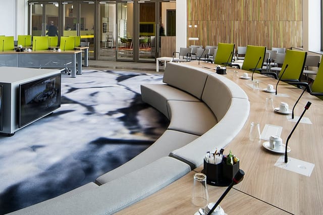 Modern kantoorruimte met beige gebogen zitbank, houten tafel, lime groene stoelen, inbouwkasten en houten wandbekleding.