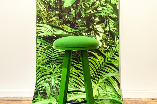 Groene barkruk met zacht zitoppervlak en drie poten, geplaatst voor een achtergrond met tropische plantenmotief.