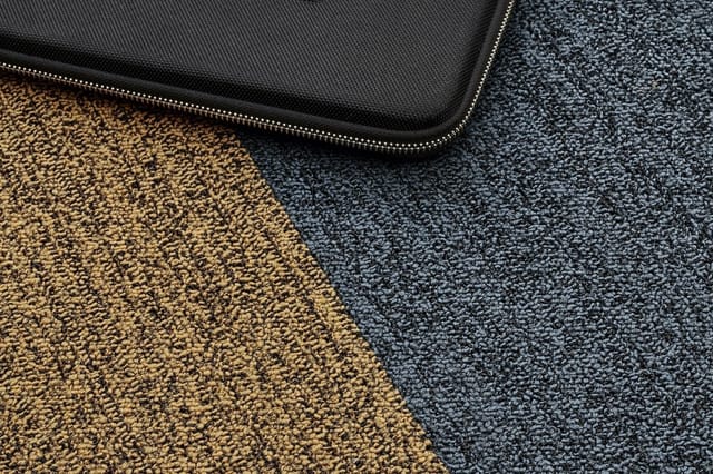 Twee tapijtstalen naast elkaar: goudkleurig en donkerblauw berber tapijt, met zwarte laptoptas met ritssluiting erboven.