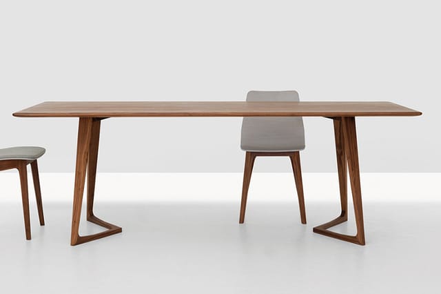 Houten vergadertafel met trapeziumvormige poten, grijze stoel en kruk in minimalistisch interieur.