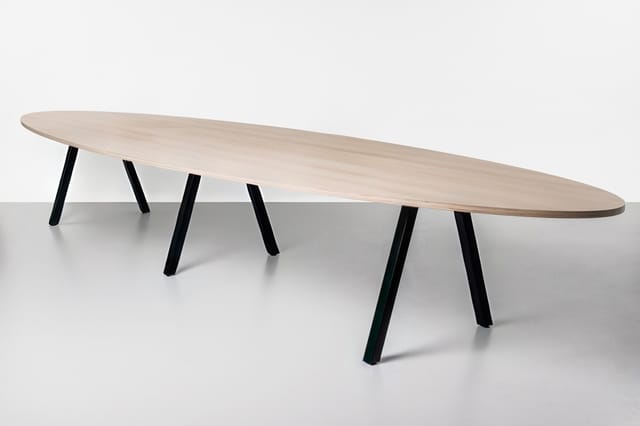 Lange ovale vergadertafel met licht houten blad en zwarte schuine poten, frontaal weergegeven.
