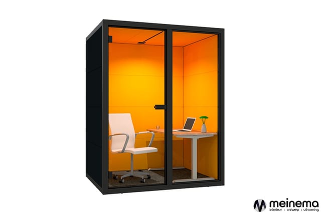 Zwarte belcel met oranje wanden, witte bureaustoel, grijs bureauoppervlak, laptop en plant erin.