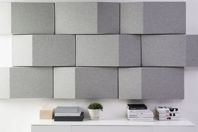 Grijze akoestische wandpanelen in verschillende tinten op witte wand boven witte plank met boeken, plant en houten kubus.