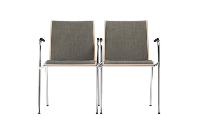 Twee grijze wachtstoelen met houten rug, chromen armleuningen en metalen poten, in tandem opgesteld tegen witte achtergrond.
